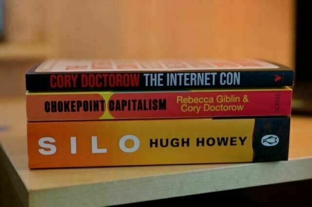 Three books piled on top of each other on a table. From the top --Cory Doctorow's The Internet Con; Chokepoint Capitalism by Rebecca Giblin and Cory Doctorow; and Silo by Hugh Howey